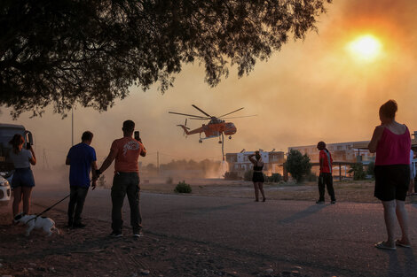 Varias personas observan un helicóptero de bomberos llenando de agua un estanque, mientras arde un incendio en el pueblo de Gennadi, en la isla de Rodas, Grecia. 