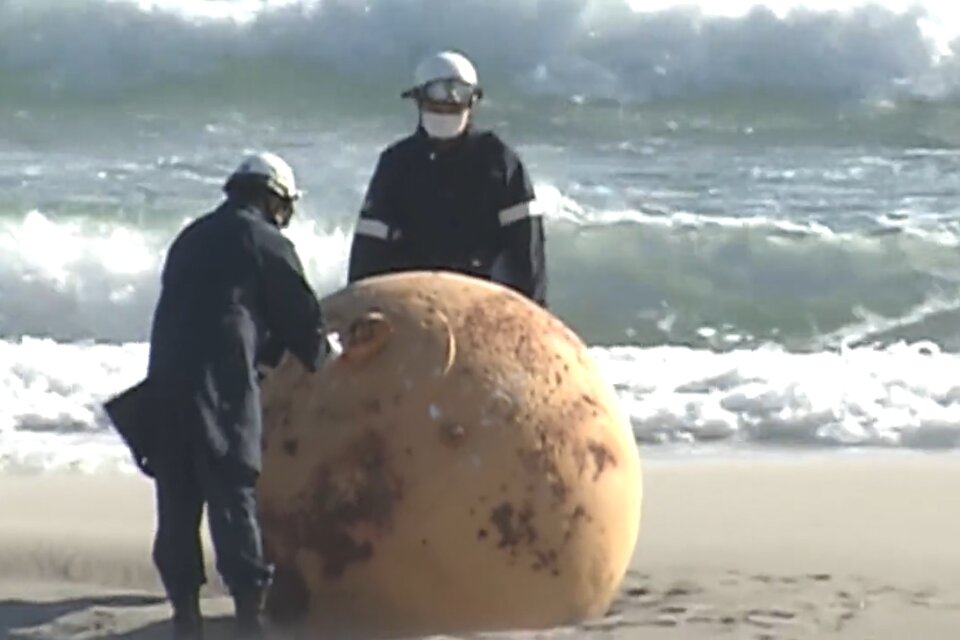 Dos agentes del escuadrón antibombas analizan la esfera de metal encontrada en la playa japonesa de Enshuhama. (Foto: captura de pantalla NHK)