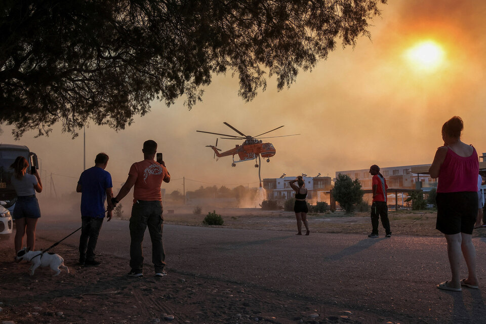 Varias personas observan un helicóptero de bomberos llenando de agua un estanque, mientras arde un incendio en el pueblo de Gennadi, en la isla de Rodas, Grecia.  (Fuente: NA)