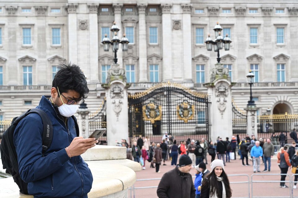 Un joven con barbijo mira su celular en la entrada a Buckingham Palace en Londres. (Fuente: AFP)